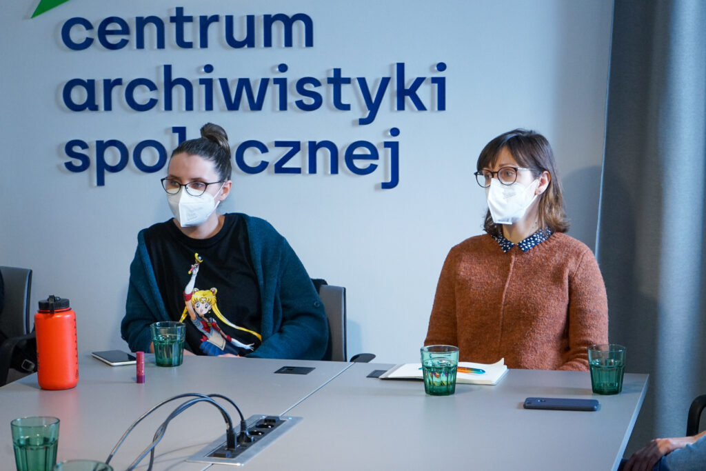 Dwie gościnie z Czeskiej Akademii Nauk siedzą w maseczkach przy stole. Za nimi logo CAS. 