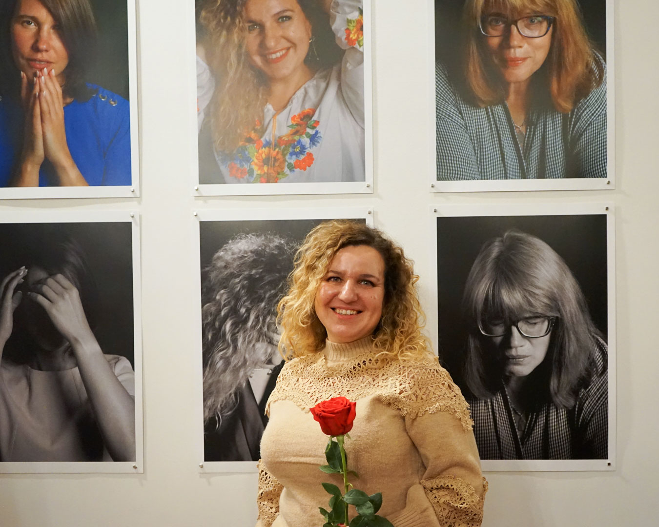 Portret kobiety w kręconych, blond włosach na tle prezentowanych na ścianie innych portretów kobiet. W dłoni kobieta trzyma czerwoną różę.